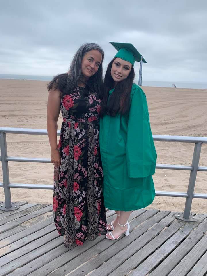 Priscilla and her daughter pose for a photo on the boardwalk of a beach in NYC. Priscilla is wearing a floral dress and her daughter is wearing a graduation cap and gown.