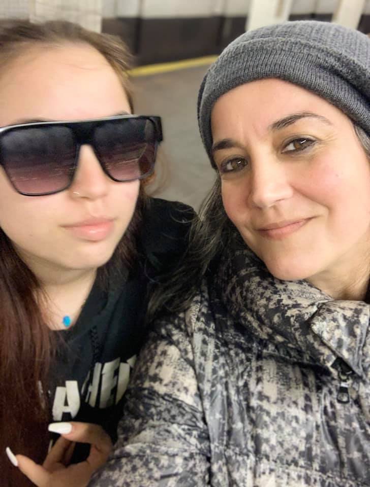 This photo shows Priscilla and her daughter taking a selfie in the NYC subway system on a chilly day. Priscilla is wearing a warm coat and a beanie. Her daughter is wearing big sunglasses.